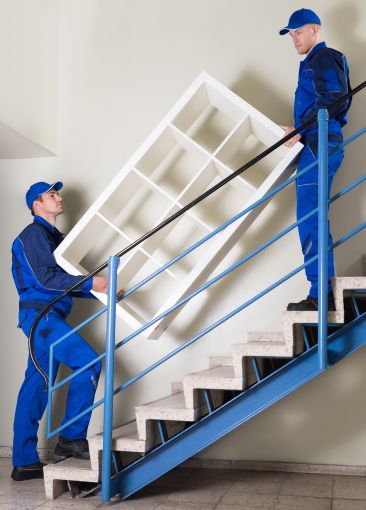 Two furniture movers carrying a white shelving unit up a staircase